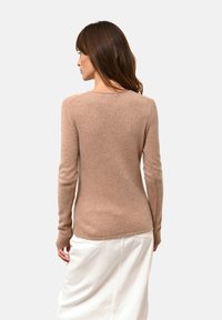 Hellbrauner, taillierter Pullover mit langen Ärmeln, weicher Textur und rundem Halsausschnitt, kombiniert mit einem weißen Rock. Nahtdetails entlang der Schultern.