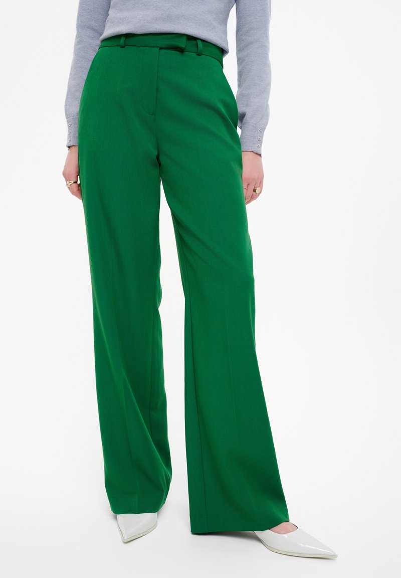 Groene wijde broek met een riem, voorzien van een gladde stoftextuur en voorplooien. Gepaard met witte schoenen met een spitse neus.