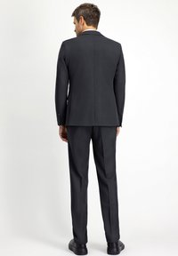 Abito nero con spalle strutturate e uno spacco posteriore, abbinato a pantaloni coordinati e scarpe nere. Tessuto liscio e formale.