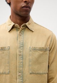 Cămașă din denim galben cu închidere prin butoni, guler ascuțit, două buzunare la piept și textură decolorată. Prezintă cusături în contrast.
