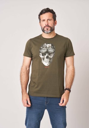 Man met baard die een olijfgroene t-shirt draagt met schedelafbeelding en tekst "Free Soul", staand tegen een effen lichte achtergrond.