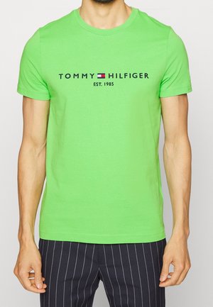 Mężczyzna ubrany w jaskrawozieloną koszulkę Tommy Hilfiger z logo i napisem "EST. 1985", w połączeniu z czarnymi spodniami z białymi pionowymi paskami.