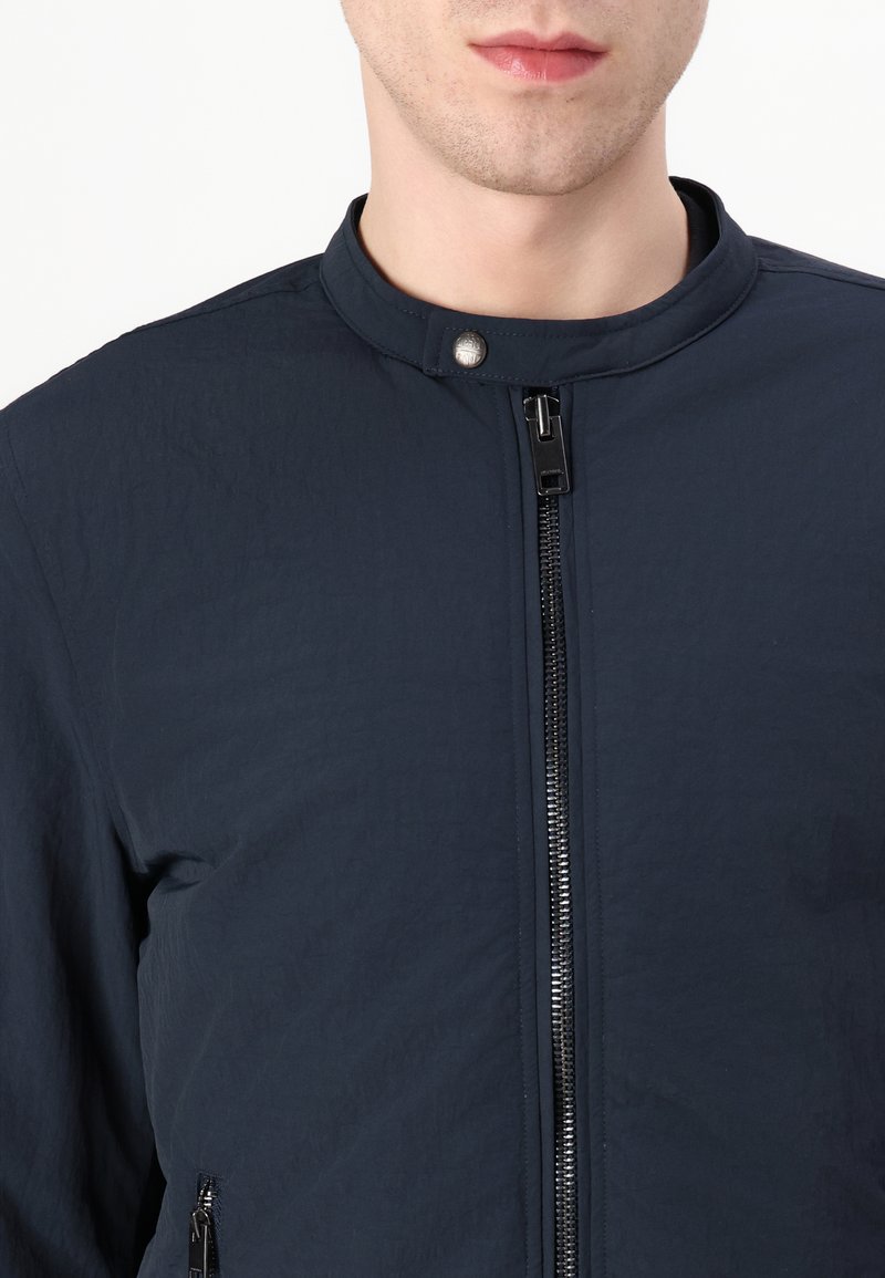 Veste légère bleu marine avec col montant, fermeture éclair à l'avant et tissu texturé. Comprend une fermeture à bouton pression et des poches latérales.