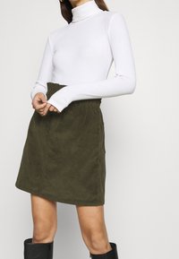 Vrouw in een witte longsleeve coltrui, olijfgroene hoge taille rok en zwarte over de knie laarzen, staande met gevouwen handen.