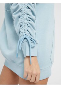 Felpa azzurro chiaro con maniche arricciate, dotata di lacci regolabili ai polsini e una texture morbida. Cuciture distintive lungo le braccia.