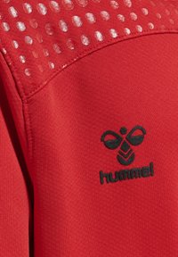 Roter Sweatshirt mit einem strukturierten oberen Teil, der kreisförmige Muster zeigt, und einem schwarzen Logo mit einem Bienenmotiv auf der linken Seite.