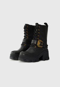 Black faux leather ankle boots with gold hardware, lace-up front, chunky heel, and strap detail with ornate buckle. Textured rubber sole.