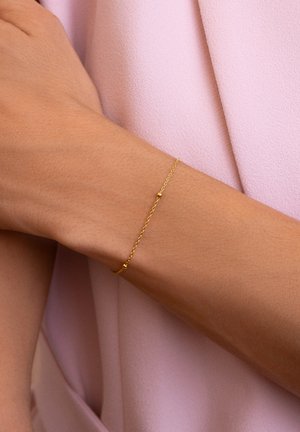 Pulsera de cadena de oro con un diseño delicado, que presenta pequeños acentos redondos y una textura retorcida. Llevada en una muñeca desnuda sobre una tela rosa.