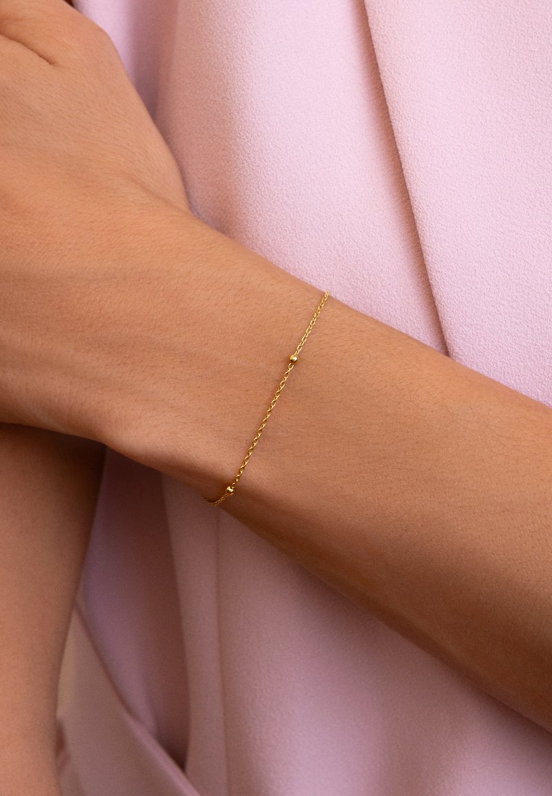 Pulsera de cadena de oro con un diseño delicado, que presenta pequeños acentos redondos y una textura retorcida. Llevada en una muñeca desnuda sobre una tela rosa.
