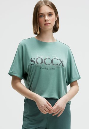 Young woman with short brown hair wears a teal T-shirt with "Keep your flow, SOCCX & Shine, Creating Stories" text, standing against a white background.