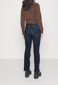 Mujer con cabello oscuro y largo, vistiendo un suéter marrón, jeans azul oscuro y zapatos de plataforma negros, de pie de espaldas sobre un fondo liso.