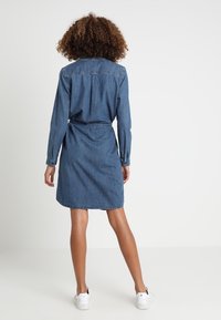 Jeansskjolklänning med lång ärm, bälte i midjan och knälång längd. Blått tyg med en slät textur. Vita sneakers.