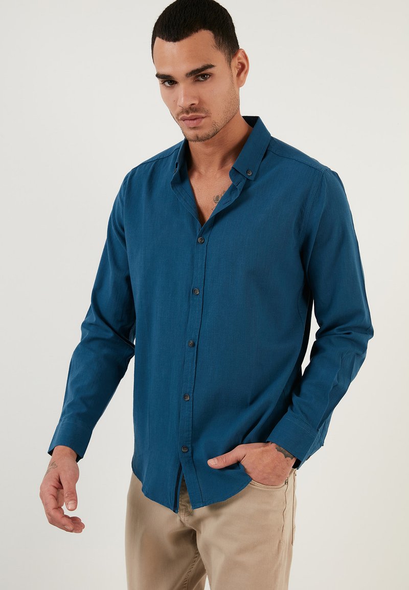 Camisa de botones color verde azulado con mangas largas, hecha de un tejido suave; presenta un collar puntiagudo y botones oscuros en la parte delantera.