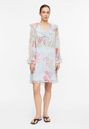 Junge Frau trägt ein hellblaues, knielanges Kleid mit Blumenmuster, Rüschenkragen und Manschetten, steht vor weißem Hintergrund.