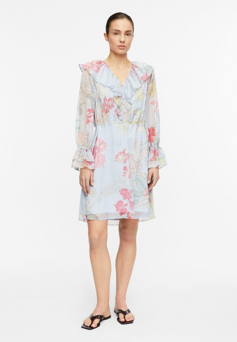 Junge Frau trägt ein hellblaues, knielanges Kleid mit Blumenmuster, Rüschenkragen und Manschetten, steht vor weißem Hintergrund.