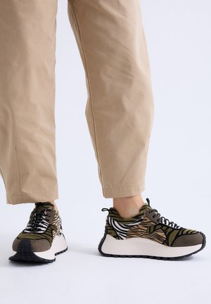 Persona con pantalones beige y zapatillas con estampado animal de rayas negras y blancas y suelas gruesas blancas sobre fondo blanco.