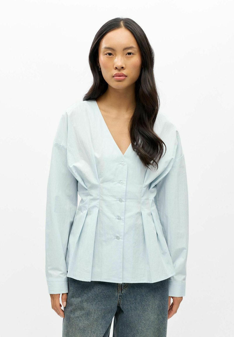 Femme aux longs cheveux foncés porte un chemisier bleu clair boutonné avec un col en V profond et des détails plissés, associé à un jean bleu.