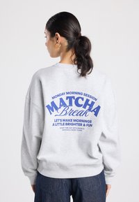 Sweat-shirt gris clair à coupe décontractée, avec un texte bleu au dos. Conçu avec un col rond et des poignets côtelés, simple et confortable.