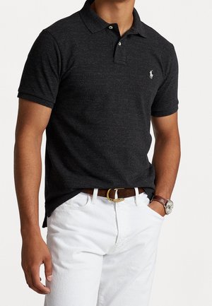 Homme portant un polo gris foncé à manches courtes, un pantalon blanc avec une ceinture marron et une montre au poignet gauche.