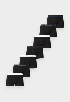 Stehender oberkörperfreier Mann mit schwarzen Tommy Hilfiger Boxershorts vor einem dunkelgrünen Ledersofa auf hellem Teppichboden.