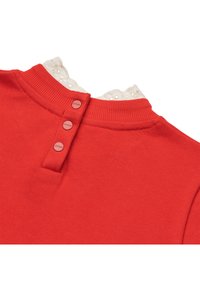 Maglione rosso con colletto a coste, caratterizzato da un bordo in pizzo bianco e tre bottoni coordinati sulla parte posteriore del collo. Texture morbida, design casual.