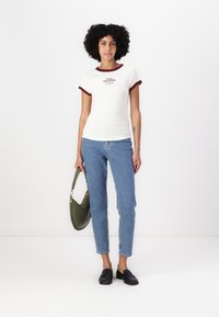 Bela majica s temno rdečim ovratnikom in rokavi, ki ima logotip. V kombinaciji s svetlo modrimi ravnimi jeans hlačami in zeleno torbico. Črne loafer čevlje.