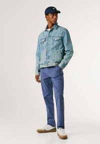 Vīrietis, kurš valkā gaiši zilu džinsa jaku, tumši zilu cepuri, zilas bikses un baltas sporta kurpes, stāv ar vienu roku kabatā pret baltu fonu.