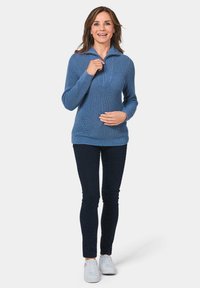 Blauer Strickpullover mit halbem Reißverschluss-Kragen, geripptem Muster, seitlichen Taschen und lockerer Passform, kombiniert mit dunklen Skinny Jeans und weißen Sneakers.