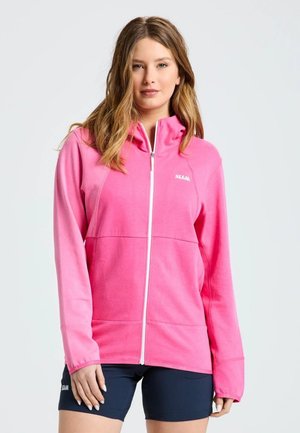 Una mujer posa con una chaqueta deportiva rosa intenso con cremallera y unos pantalones cortos de ciclismo azul marino, sobre un fondo claro y liso.