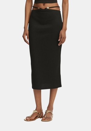 Donna che indossa una gonna midi nera a coste, cintura a catena color oro con charms e sandali piatti metallici dorati, in piedi con le braccia rilassate.