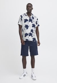Camicia a maniche corte con bottoni, su sfondo bianco e motivo floreale blu scuro. Abbinata a pantaloni corti blu e sneakers bianche.