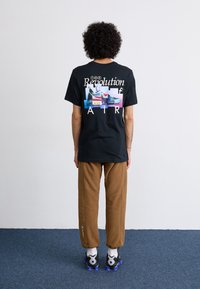 Black cotton t-shirt featuring colorful graphic text and sneaker prints on the back. Paired with brown sweatpants and black sneakers.