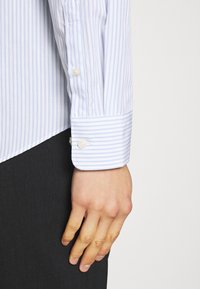 Camicia a righe azzurro chiaro e bianca con polsini abbottonati. Il tessuto appare liscio; la camicia ha un taglio su misura e maniche lunghe.