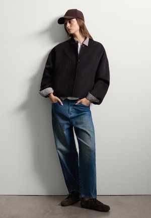 Zwarte cropped jas met een knoopsluiting, gedragen over een geruit shirt. Blauwe rechte jeans en donkerbruine sneakers maken de outfit compleet.