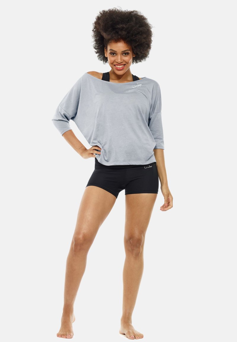 Lichtgrijze losse top met driekwart mouwen en schouderuitsnijding, gecombineerd met zwarte shorts. Soepele textuur; minimale branding.