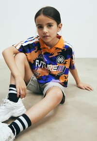 Lühikese varrukaga polo särk mitmevärvilises kamoflaažis, millel on oranž krae, logod ja lahtine istuvus, koos teksashortside ja jooksujaladega.
