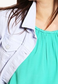 Camisa de botón gris claro superpuesta sobre una blusa turquesa texturizada con un escote fruncido. Mangas cortas y sutiles detalles de desgaste en la camisa.