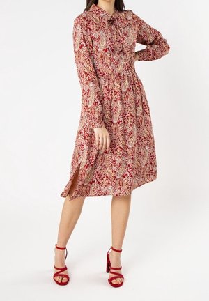 Femme portant une robe rouge à manches longues et longueur genou avec motif cachemire, fente latérale, et sandales rouges à talons bloque et brides, posant avec une main sur la hanche.