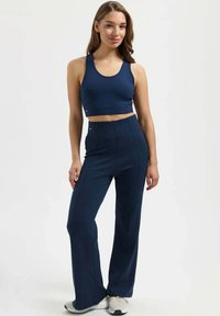 Nuori nainen, jolla on yllään tummansininen hihaton vartaloa myötäilevä toppi ja vyötäröltä korkeavyötäröiset leveälahkeiset housut, seisoo käsi lantiolla ja hymyilee.