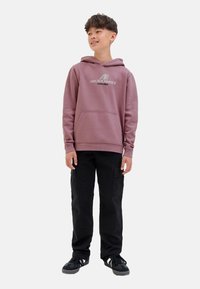 Un jeune souriant se tient debout, portant un sweat à capuche rose avec logo, un pantalon cargo noir et des baskets noires, contre un fond blanc uni.