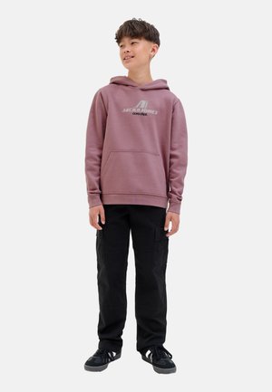 Un jeune souriant se tient debout, portant un sweat à capuche rose avec logo, un pantalon cargo noir et des baskets noires, contre un fond blanc uni.