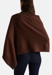 Femme portant un châle en tricot marron foncé drapé sur les épaules, associé à un jean bleu foncé, se tenant devant un fond clair uni.