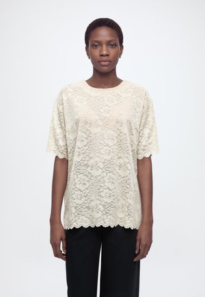 Kvinde stående og vendt fremad iført en cremefarvet bluse med korte ærmer i blonde og sorte bukser mod en simpel hvid baggrund.