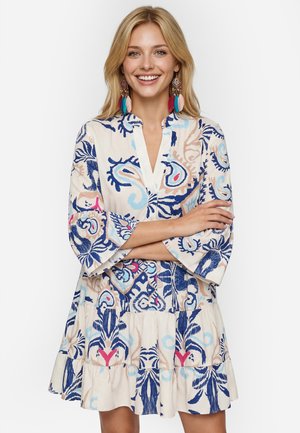 Femme blonde souriante, bras croisés, portant une robe colorée à motif cachemire et de grandes boucles d'oreilles en plumes multicolores.