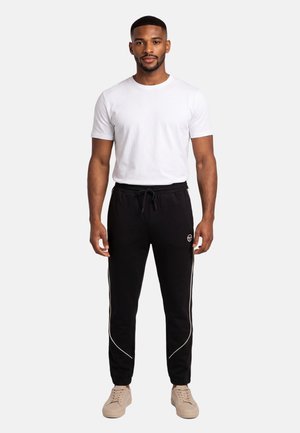 Homme debout face à l'avant, portant un t-shirt blanc uni, un jogging noir avec cordon de serrage et passepoil blanc, ainsi que des baskets beiges sur fond blanc.