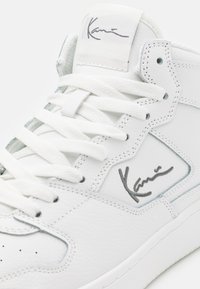 Hvit høy toppen sneaker i teksturert lær med flate snørebånd, med en signatur på siden og en etikett på tungen.