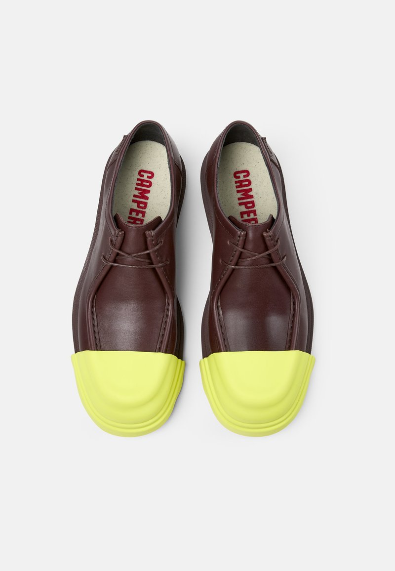 Brązowe skórzane buty z żółtą gumową osłoną na palce. Sznurowadła widoczne na górze, a na wkładce jest napis "CAMPPER" w kolorze czerwonym.