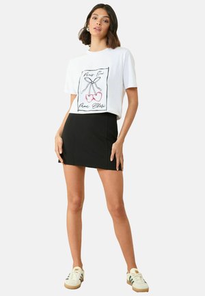 T-shirt blanc court avec un graphisme de cerise noir et rouge ; associé à une mini-jupe noire ajustée et des baskets blanches avec des accents verts.