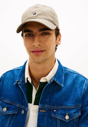 Șapcă bej cu logo mic, purtată cu o jachetă denim albastră peste o cămașă cu guler verde și alb. Textură netedă, design casual.