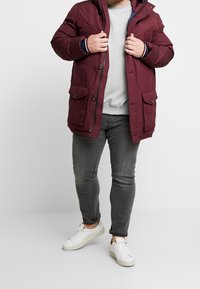 Parka color amaranto con cappuccio, dotata di due tasche frontali, chiusura con zip e texture trapuntata. Indossata sopra un maglione grigio e jeans grigi.